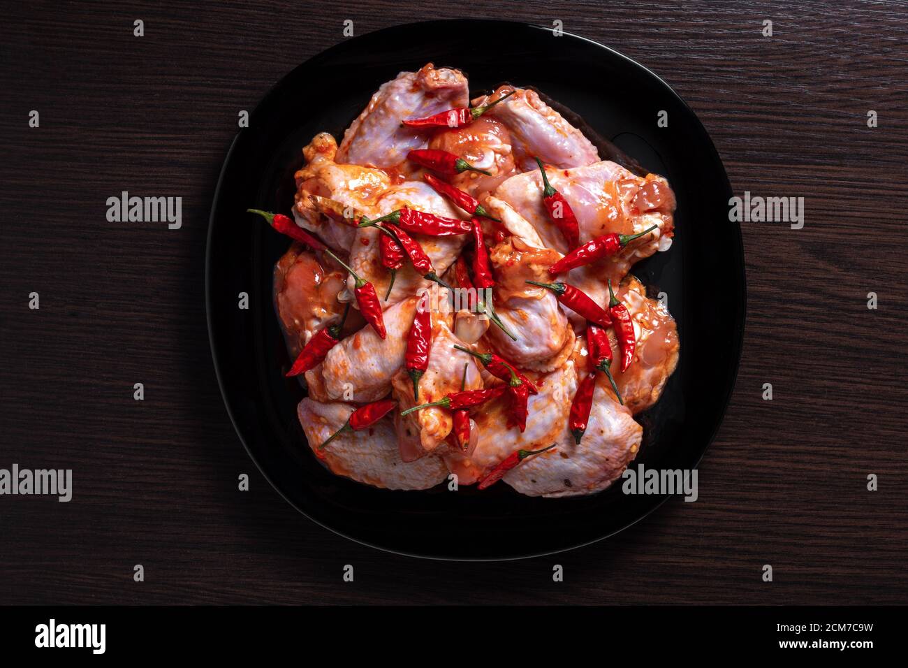 Ali di pollo crudo in una marinata con spezie su un piatto nero, un sacco di peperone rosso caldo, su un tavolo di legno scuro, vista dall'alto, concetto di cibo Foto Stock