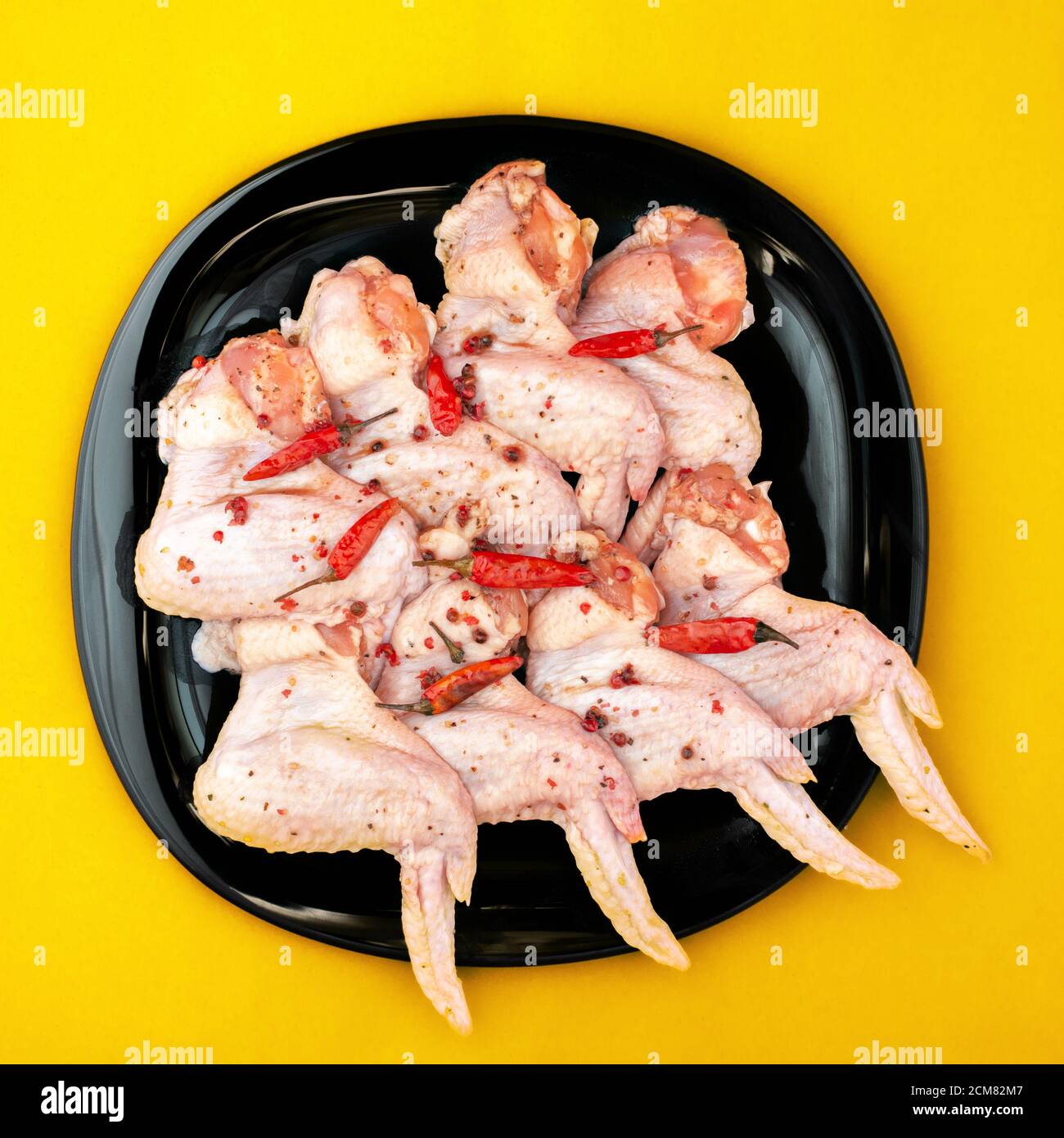 Ali di pollo crudo con spezie. Pollo crudo sottaceto con pepe caldo, su piatto nero, fondo giallo brillante Foto Stock