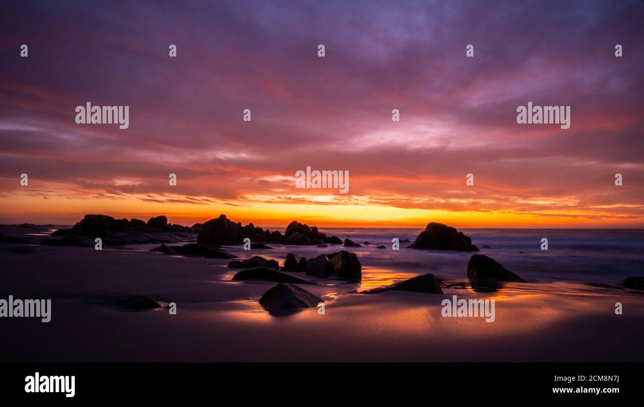 Lunga esposizione del tramonto costiero, dove i colori si riflettono nella sabbia bagnata con rocce arrotondate sullo sfondo Foto Stock