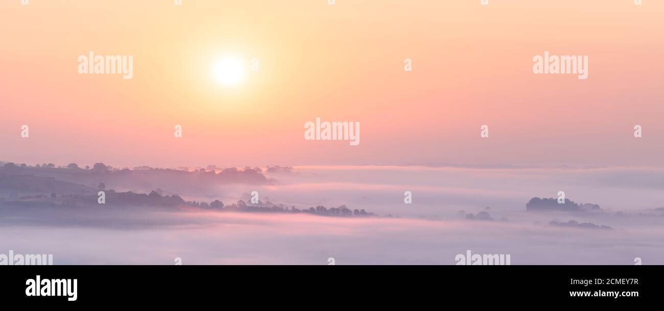 Il sole che sorge diffonde toni pastello attraverso il paesaggio velato di Lower Wharfedale, con colline alberate che svettano attraverso la nebbia. Foto Stock