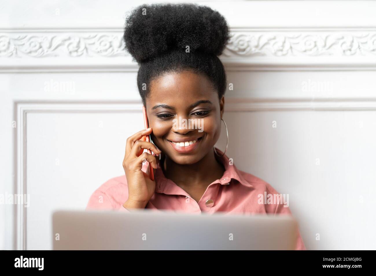 Primo piano ritratto della millenaria afroamericana con acconciatura afro in camicia rosa sorridente, parlando al telefono, guardando il computer portatile. Dipendente femminile Foto Stock