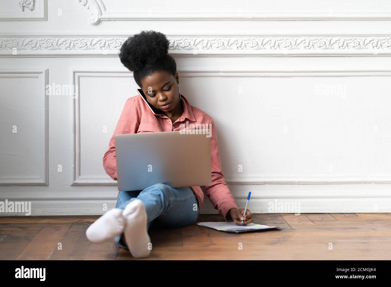 Donna afro-americana focalizzata che prende appunti, chiarisce le informazioni, parlando al telefono, utilizzando il laptop e seduto sul pavimento. Contro del dipendente biracial femminile Foto Stock