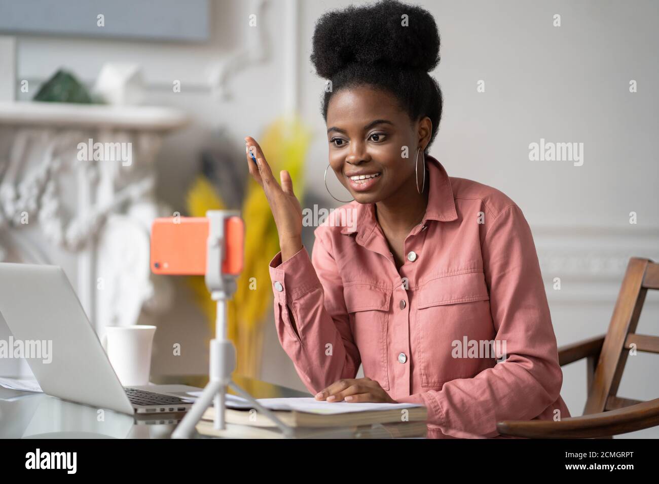 Donna millennial afroamericana con studio remoto afro-acconciatura, lavorando online su laptop, chattando con gli amici tramite videochiamata su smartphone Foto Stock
