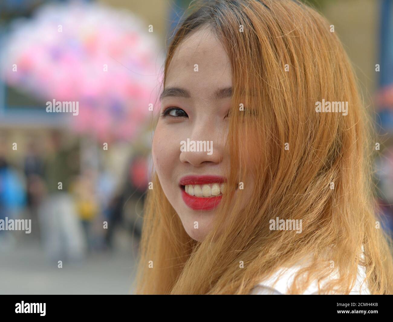 Giovane donna vietnamita con capelli tinti e rossetto rosso guarda sopra la spalla e sorride per la fotocamera. Foto Stock