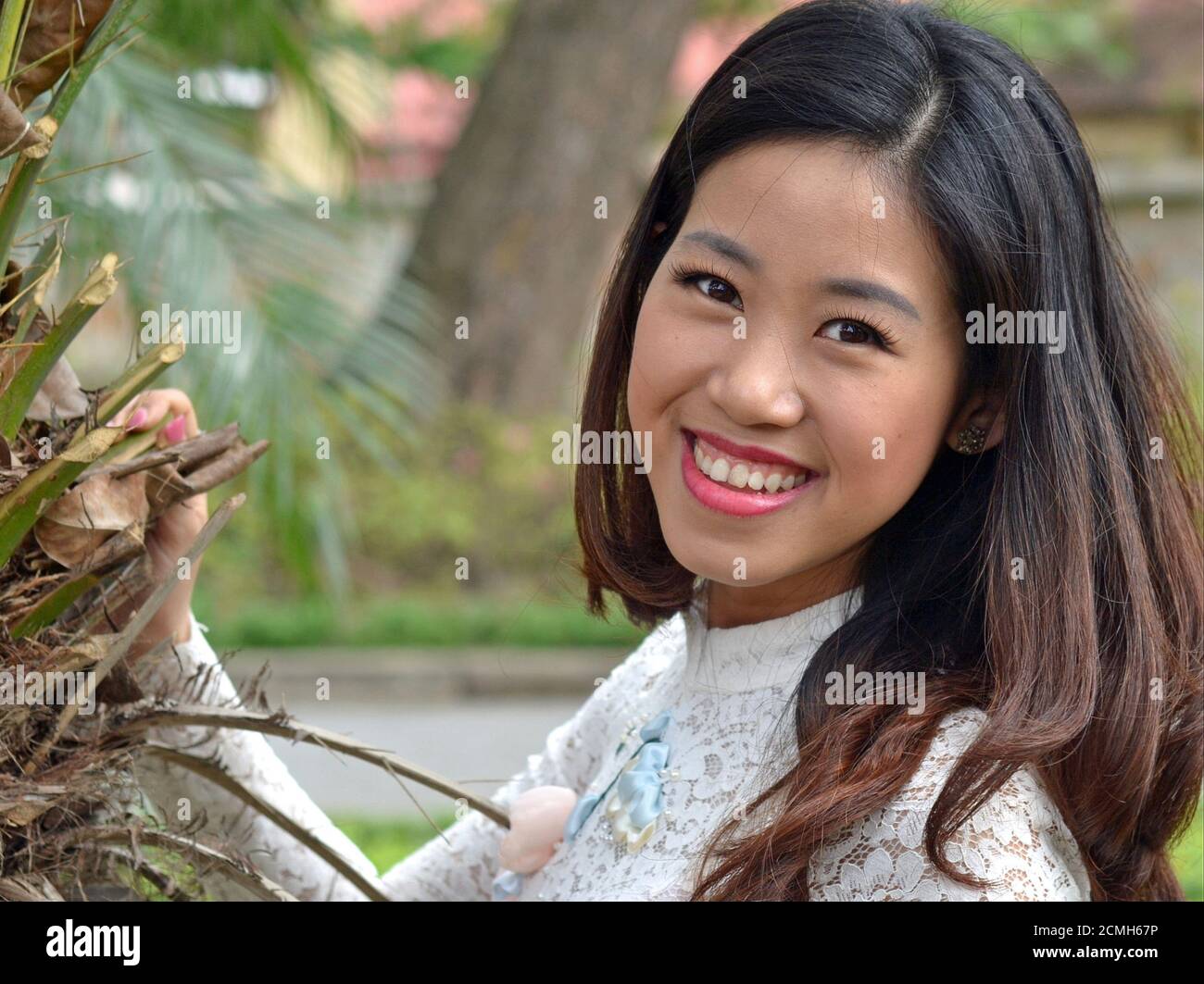 Allegra ed elegante giovane donna vietnamita con bei capelli lunghi indossa un abito in pizzo bianco e sorrisi per la macchina fotografica. Foto Stock