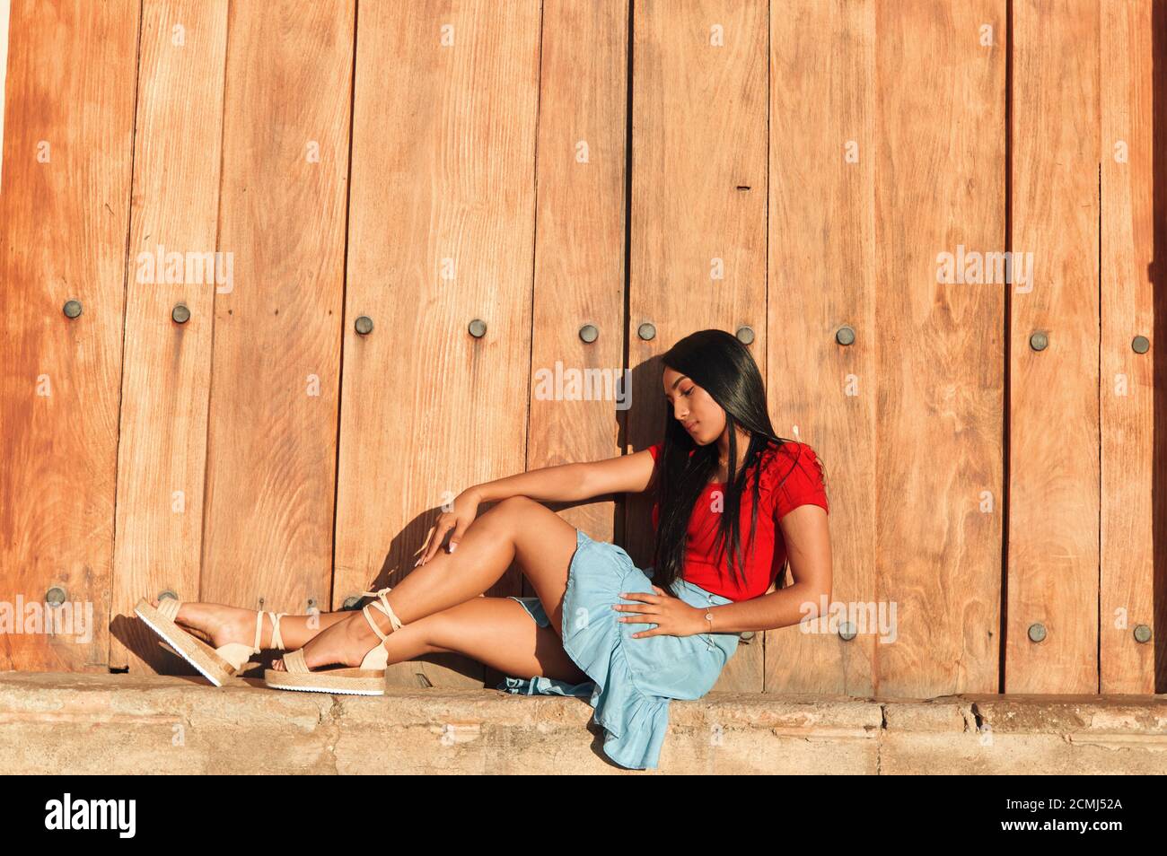 Giovane donna in una giornata estiva che indossa una camicia rossa, gonna blu brillante e sandali beige a espadrilla in posa accanto a una grande porta vintage Foto Stock