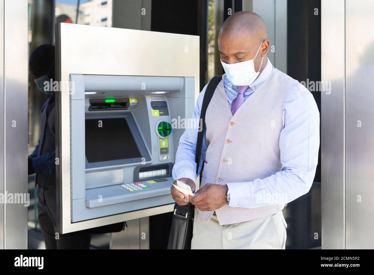 Imprenditore afro-americano che ritira denaro in un bancomat. Indossa una maschera medica come misura protettiva durante la pandemia di Covid-19. Foto Stock