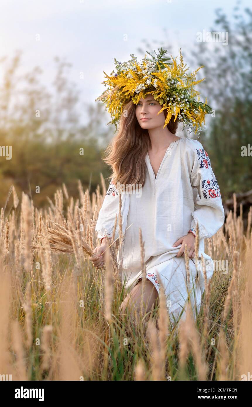 Giovane bella donna in una corona di fiori selvatici e antica camicia ricamata in un prato di erba secca gialla. Cultura Ucraina, stile etno Foto Stock