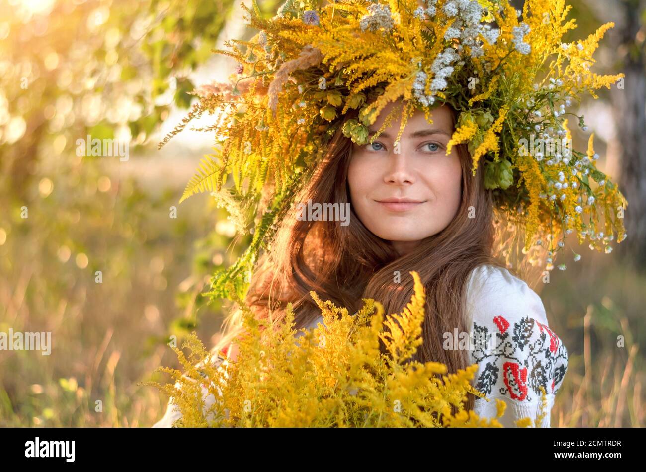 Giovane bella donna in una corona di fiori selvatici e camicia ricamata all'aperto in una giornata di sole. Primo piano verticale Foto Stock