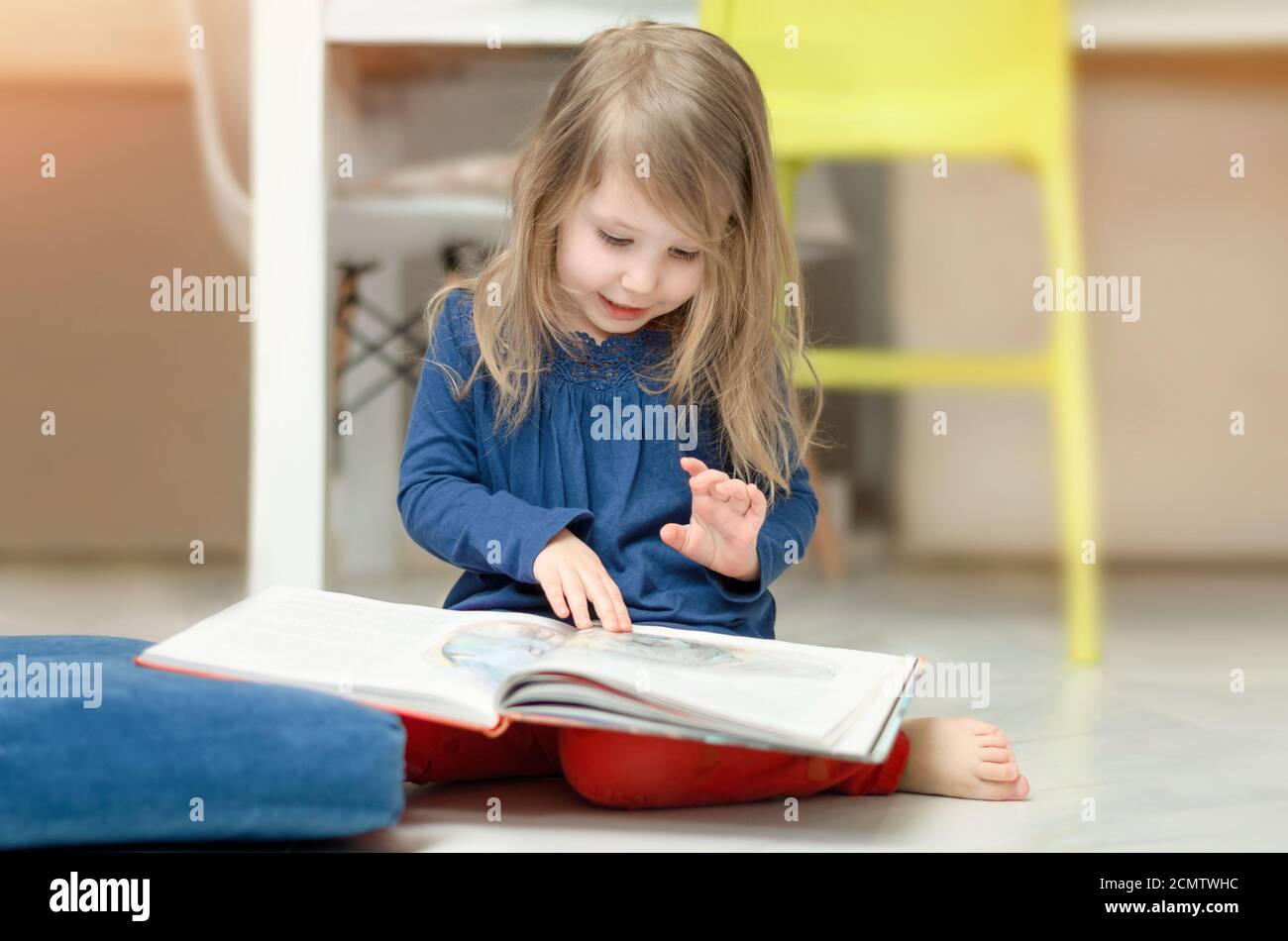 La bambina dai capelli giusti esamina le illustrazioni in un libro dei bambini seduto sul pavimento a casa. Sviluppo della prima infanzia Foto Stock
