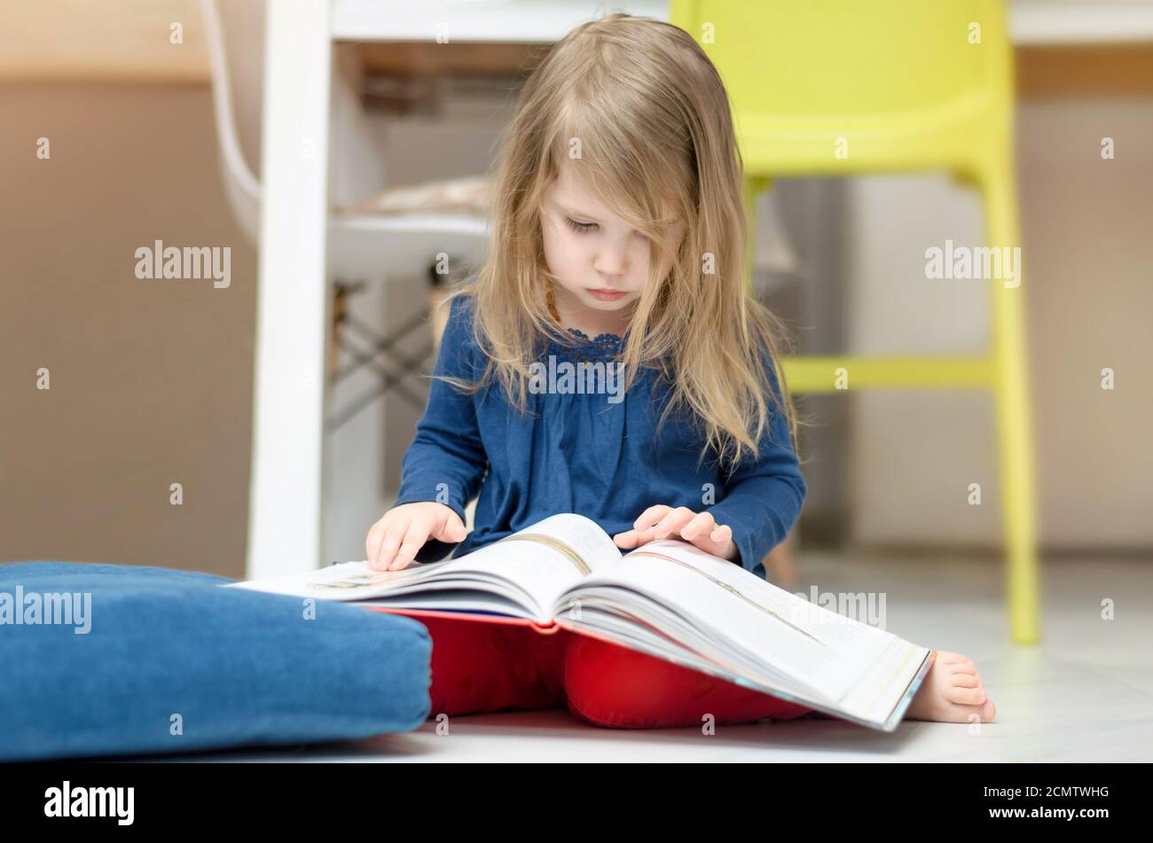 La bambina dai capelli giusti sta leggendo un libro mentre si siede sul pavimento a casa. Istruzione della prima infanzia Foto Stock