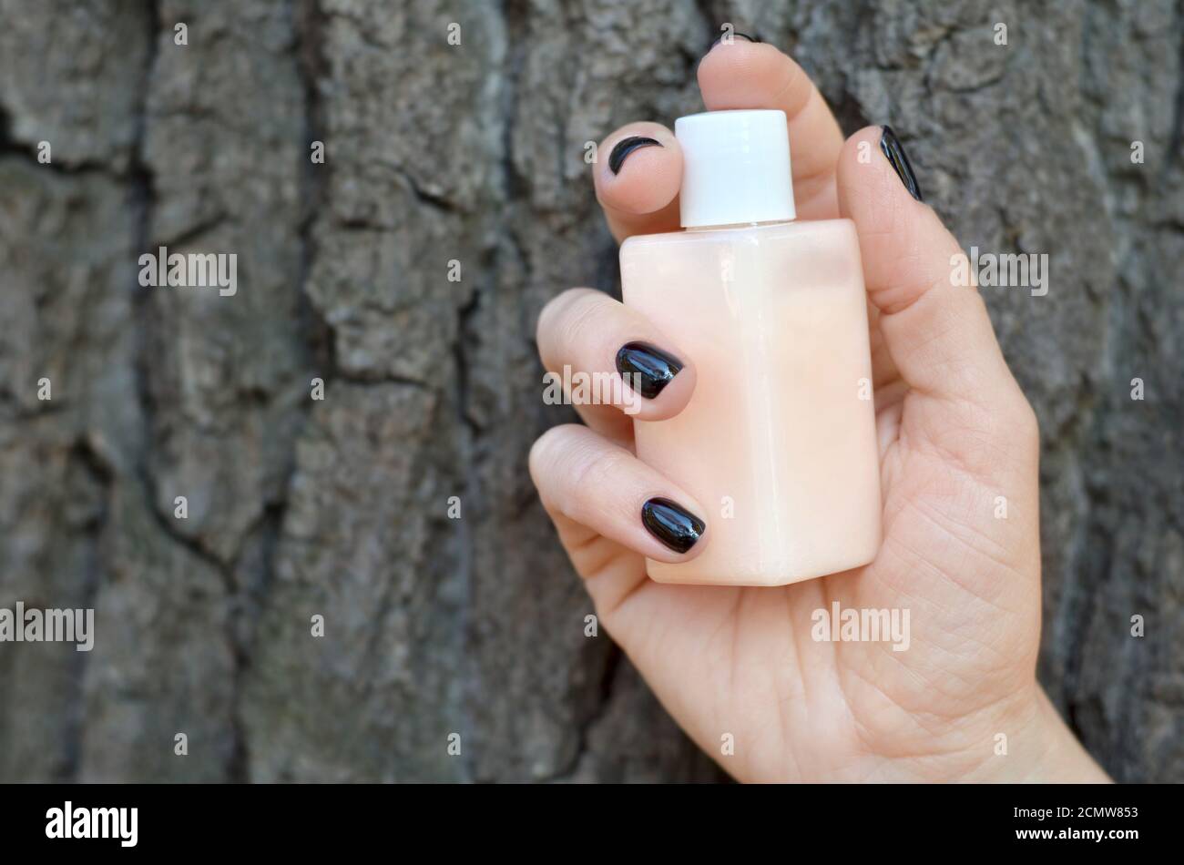 La mano della donna con unghie nere tiene una bottiglia di cosmetici naturali sullo sfondo della corteccia dell'albero. Prodotto cosmetico a base di componenti naturali Foto Stock