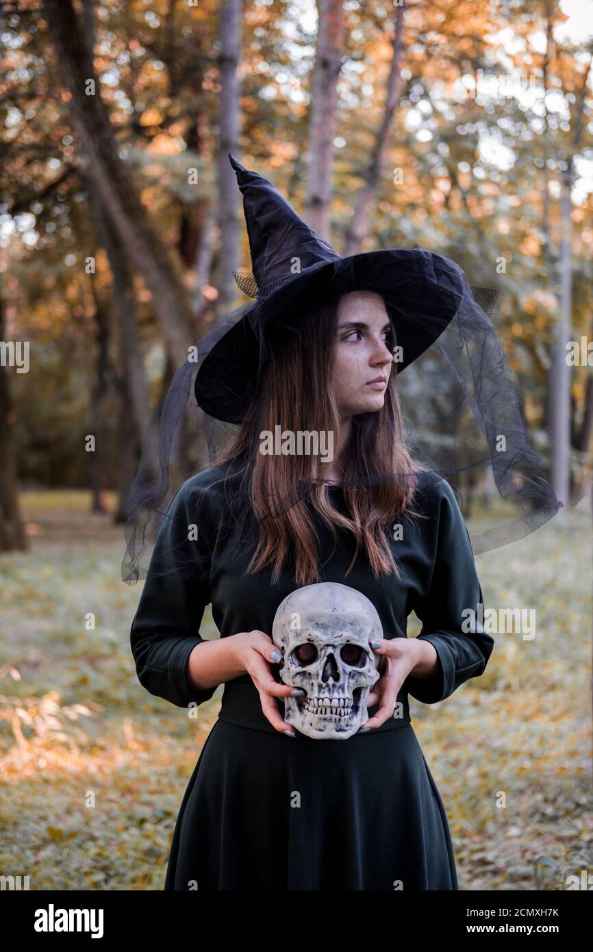 Giovane bella donna carina in abito scuro e cappello di strega tiene un cranio nelle sue mani. Costume da festa di Halloween. Foresta, parco con alberi d'autunno Foto Stock