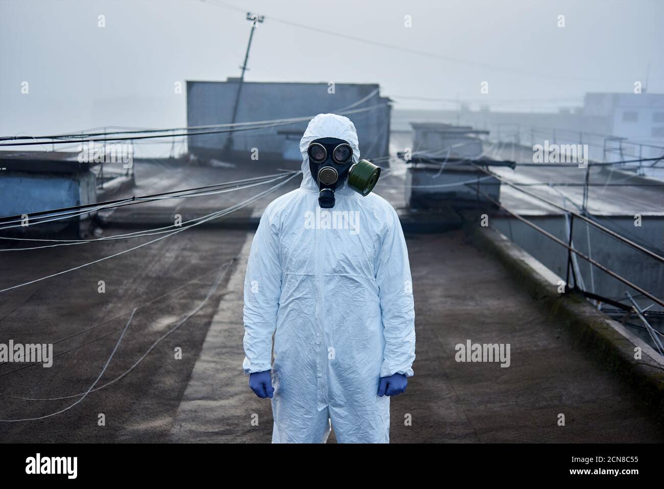 Ricercatore in piedi sul tetto di un edificio di alto piano che indossa tuta di protezione bianca e maschera a gas guardando la telecamera Foto Stock