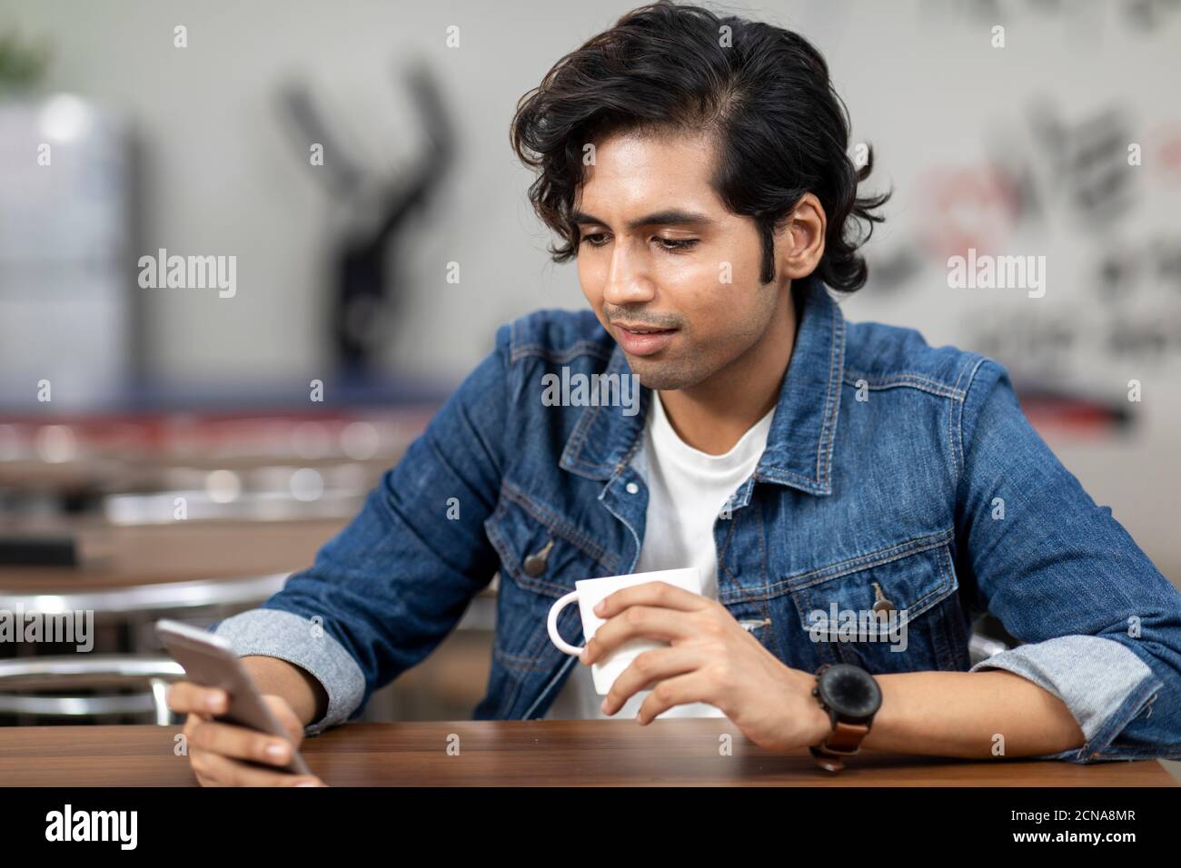 Ritratto di un giovane bell'indiano che controlla il suo telefono, seduto in una caffetteria d'ufficio, in una caffetteria, in un ambiente di lavoro informale. Foto Stock