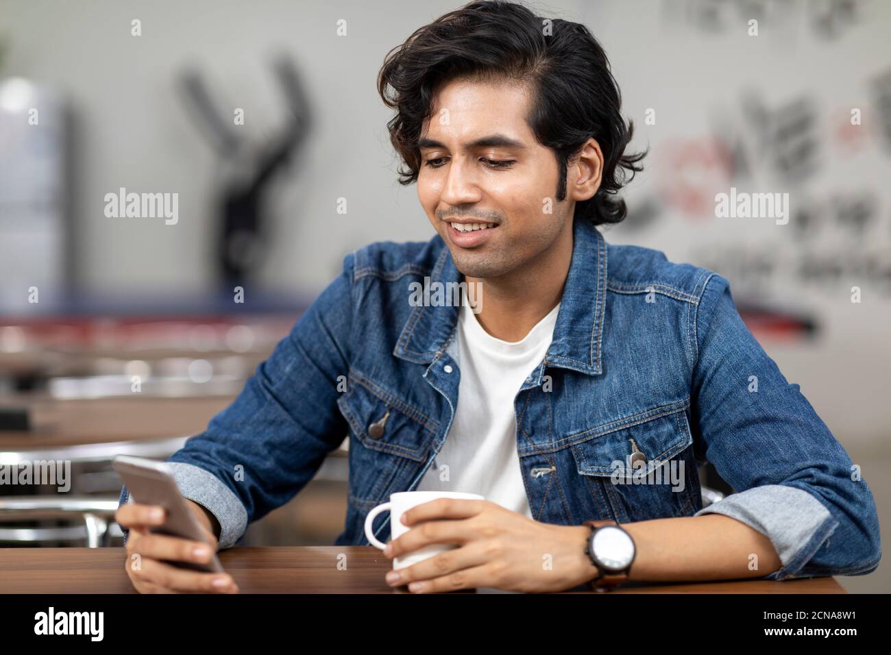 Ritratto di un giovane bell'indiano che controlla il suo telefono, seduto in una caffetteria d'ufficio, in una caffetteria, in un ambiente di lavoro informale. Foto Stock