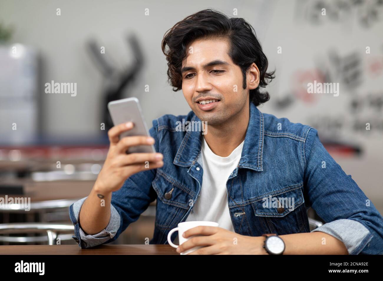 Ritratto di un giovane bell'indiano che controlla il suo telefono, seduto in una caffetteria d'ufficio, in una caffetteria, in un ambiente di lavoro informale. Foto Stock