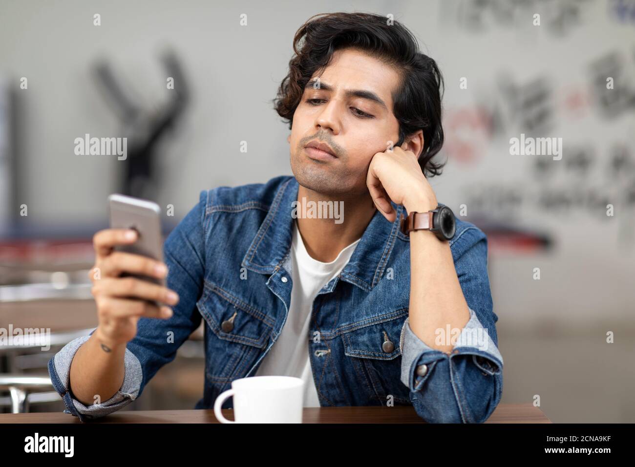 Ritratto di un giovane bell'indiano che controlla il suo telefono, seduto in una caffetteria d'ufficio, in una caffetteria, in un ambiente di lavoro informale. Foto Stock
