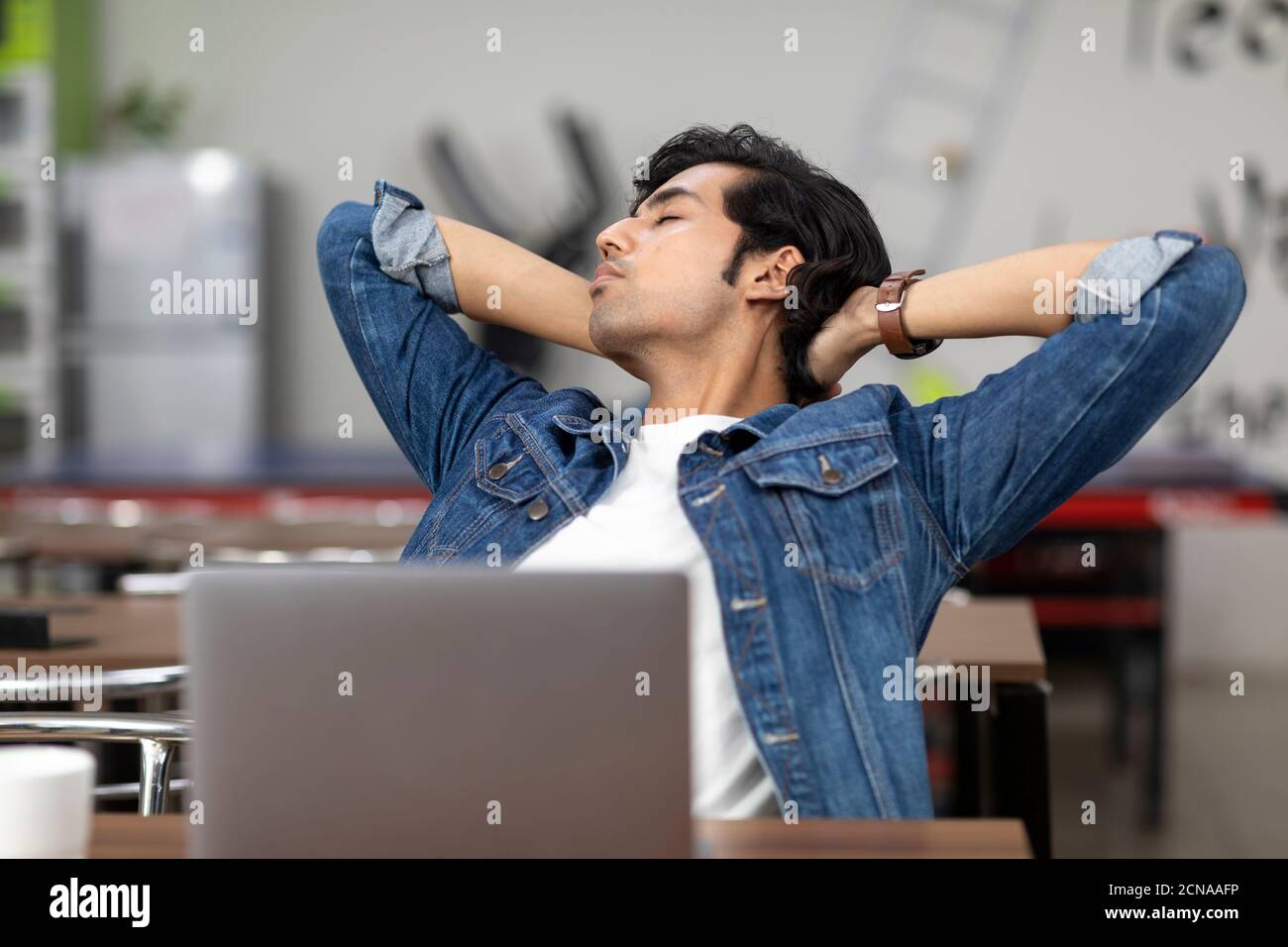 Un uomo indiano stanco che lavora sul suo computer portatile, seduto in una caffetteria d'ufficio, in una caffetteria, in un ambiente di lavoro informale, gli occhi chiusi e riposanti. Foto Stock