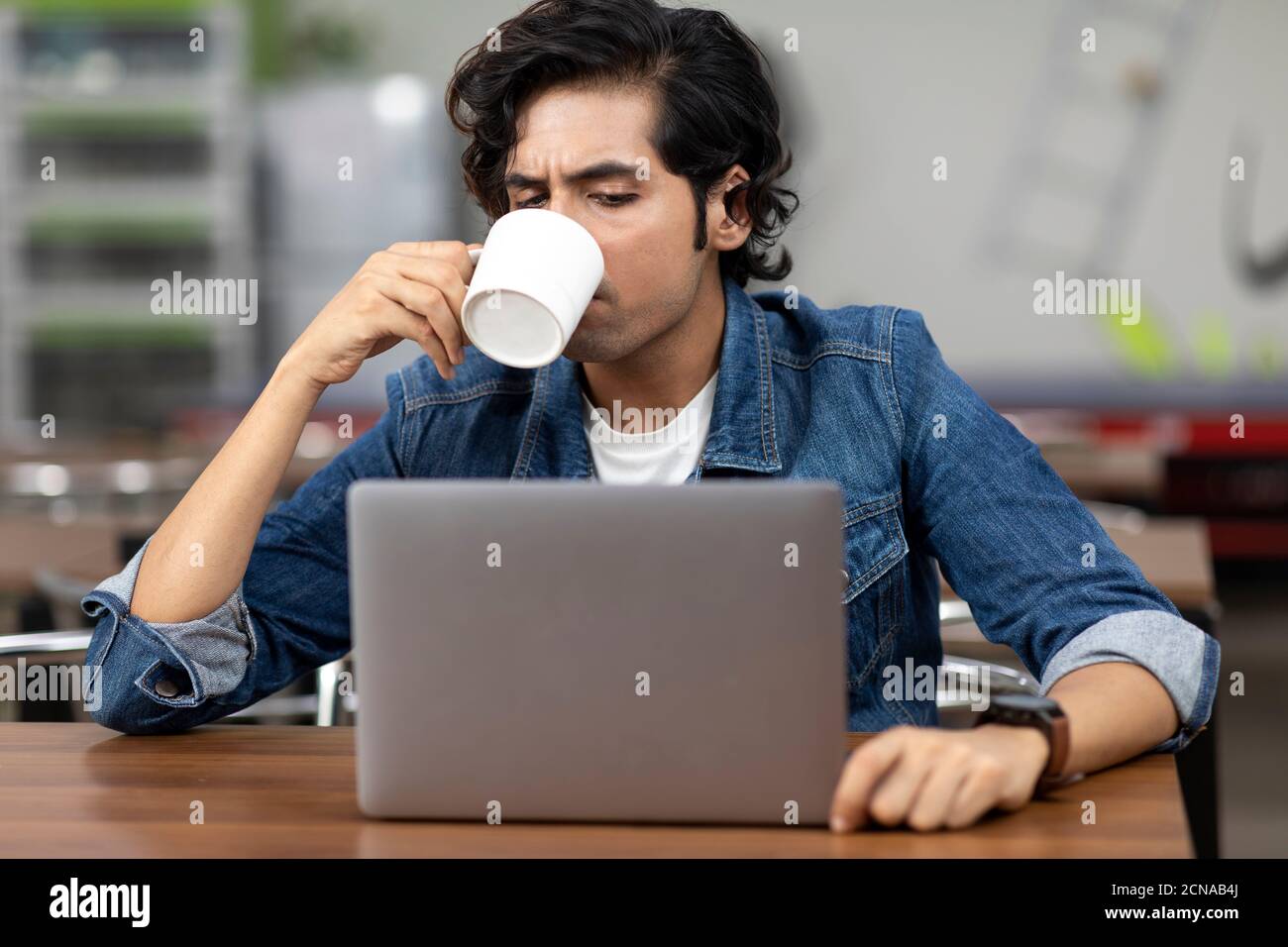 Ritratto di un giovane bell'indiano che sorseggia da una tazza di caffè, seduto in una caffetteria d'ufficio, in una caffetteria, in un ambiente di lavoro informale. Foto Stock