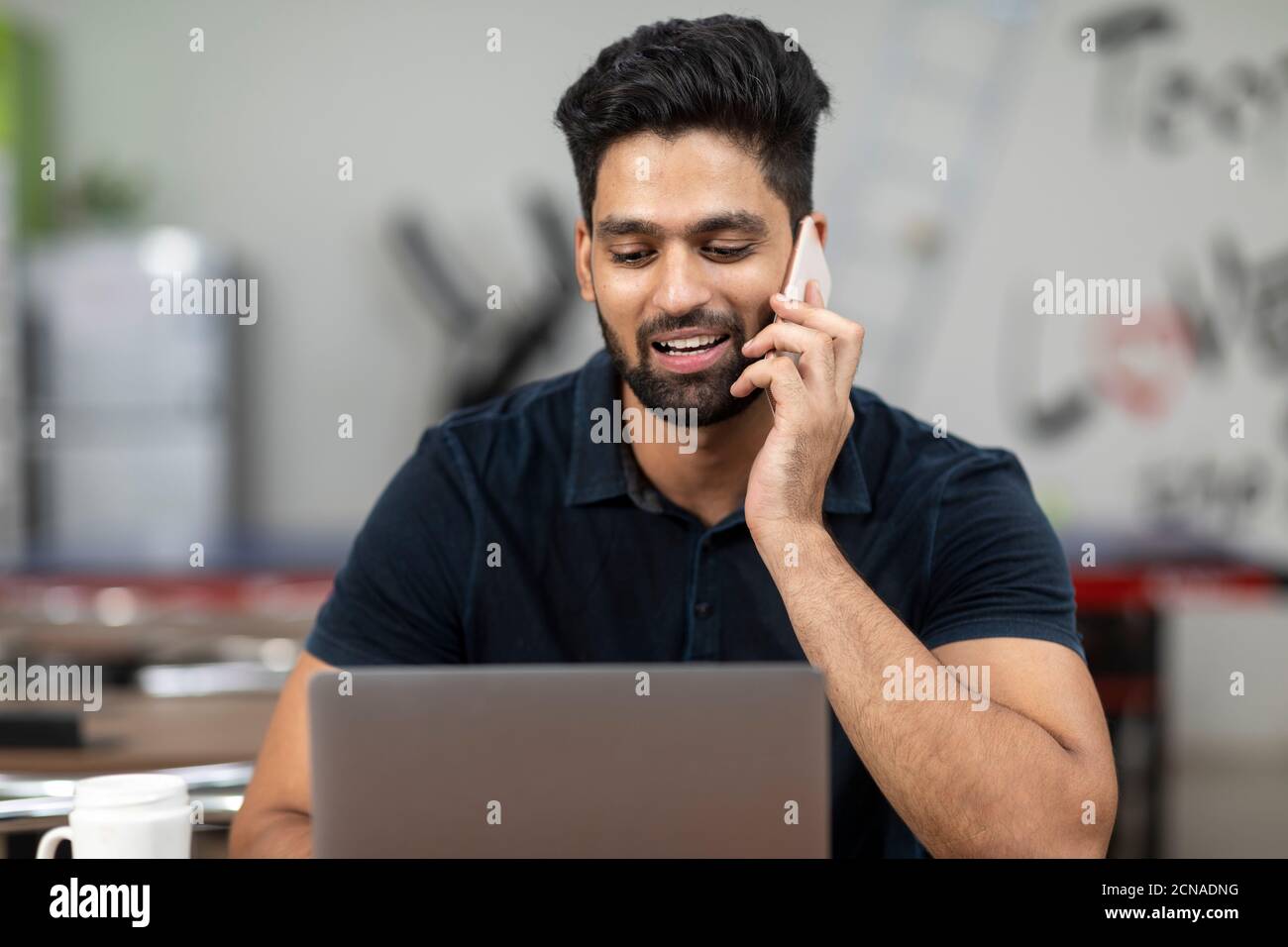 Ritratto di un giovane bell'indiano che parla al suo telefono e lavora sul suo laptop, seduto in una caffetteria d'ufficio, in una caffetteria, in un lavoro informale. Foto Stock