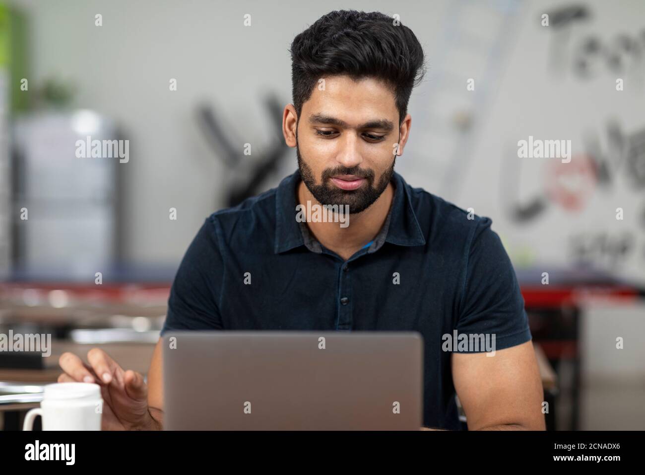 Ritratto di un giovane bell'indiano che lavora sul suo portatile, seduto in una caffetteria d'ufficio, in una caffetteria, in un ambiente di lavoro informale. Foto Stock