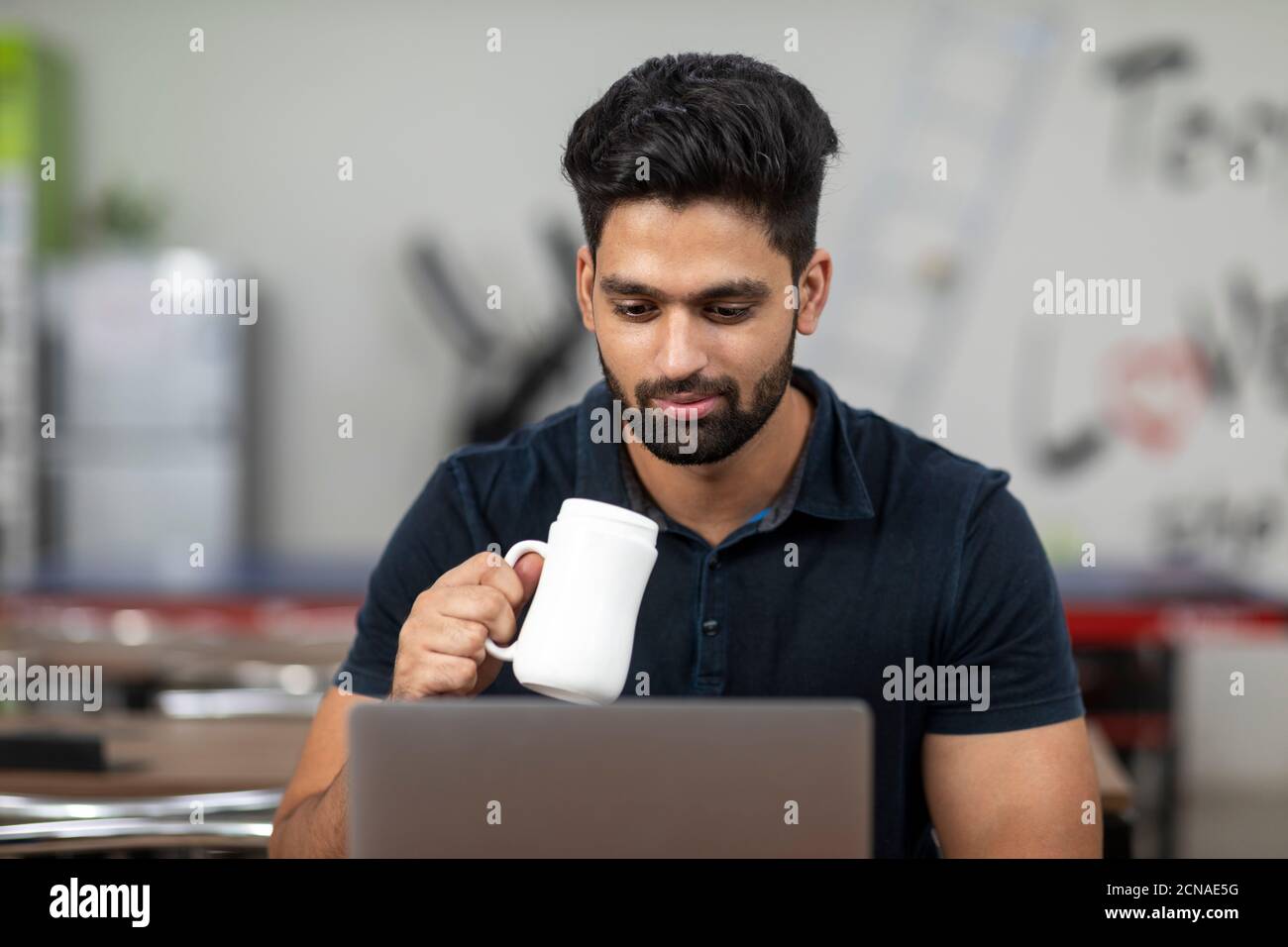 Ritratto di un giovane bell'indiano che tiene una tazza da caffè e lavora sul suo computer portatile, seduto in una caffetteria d'ufficio, in una caffetteria, in un lavoro informale. Foto Stock