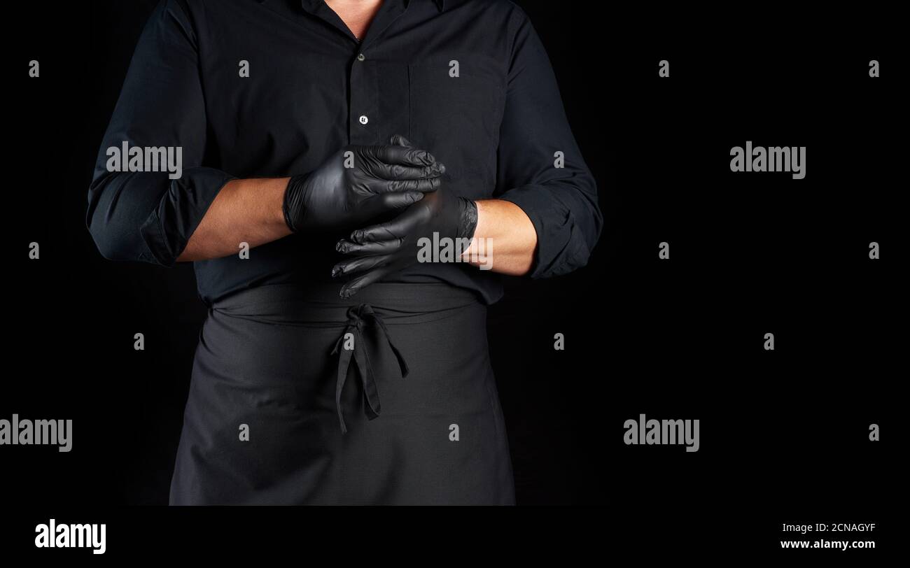chef in camicia nera e grembiule mette guanti in lattice nero sulle sue mani prima di preparare il cibo Foto Stock