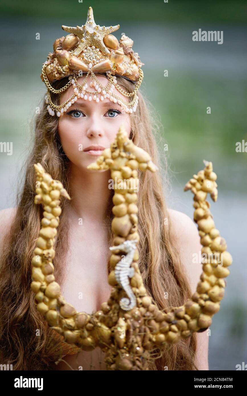 Fantasy donna vera sirena con tridente mito dea del mare con coda dorata  seduta al tramonto su rocce. Oro conchiglie corona gioielli perle. Merma  Foto stock - Alamy