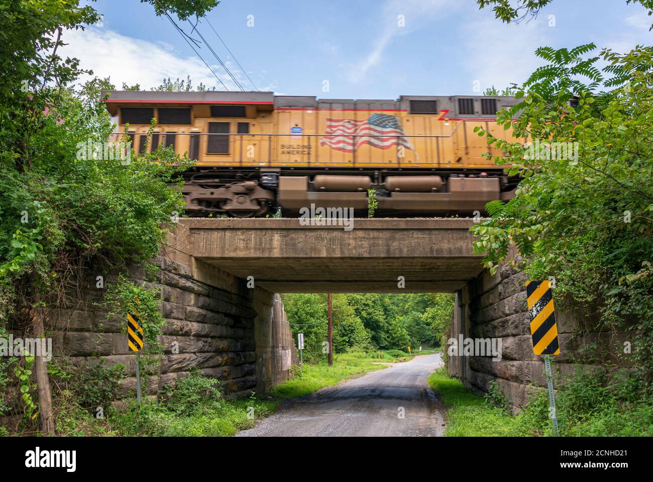 Un motore ferroviario arancione attraversa un cavalcavia in cemento e pietra e una strada solitaria in ghiaia. Foto Stock