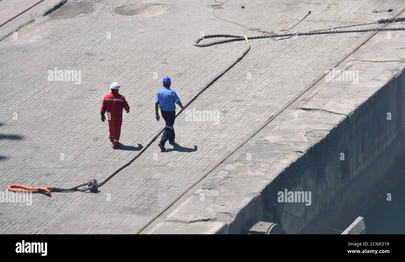 Due stevedores o uomini ormeggiatori in stoffa da lavoro e caschi di sicurezza che camminano e si avvicinano alla corda di ormeggio della nave in arrivo per fissare la linea. Foto Stock