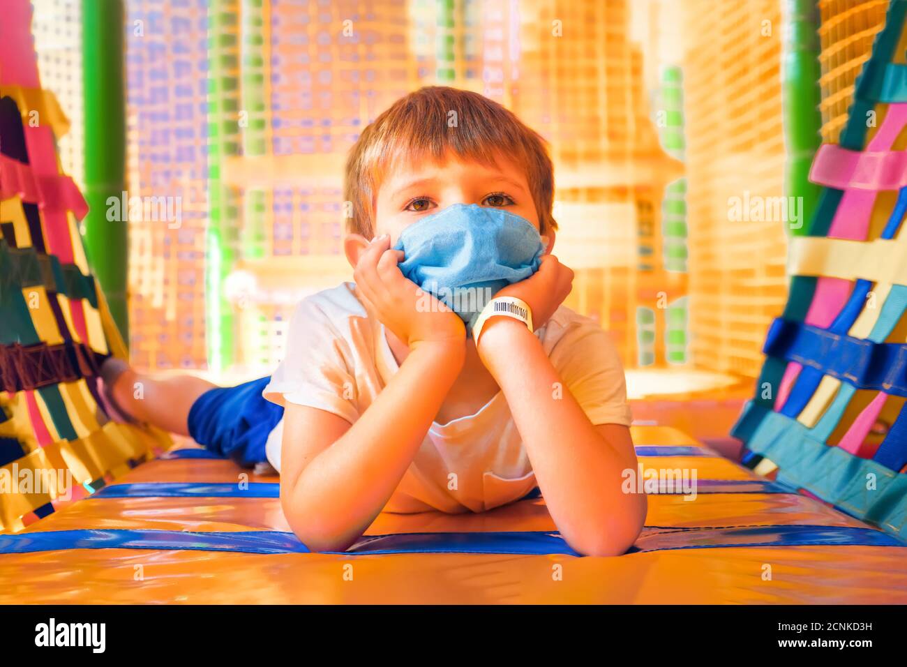 Scolaro in maschera protettiva giacente sul pavimento in sala giochi. Scuola elementare durante la quarantena. Ragazzo allegro in maschera in asilo. Sociale Foto Stock