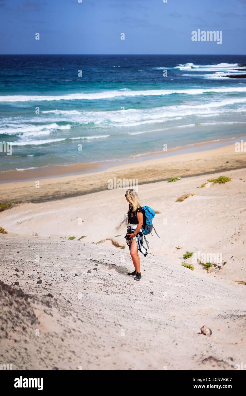 Donna escursionista con zaino scendendo una duna di sabbia verso la spiaggia solitaria. Sao Vicente Capo Verde. Foto Stock