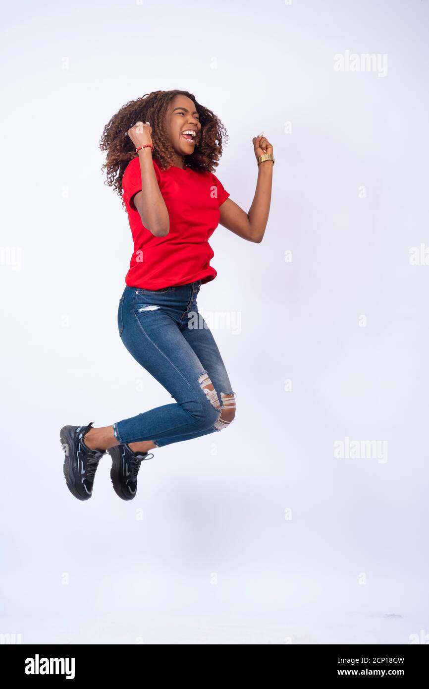 bella giovane donna africana che indossa top rosso e jeans blu che saltano con eccitazione e felicità, su sfondo bianco Foto Stock
