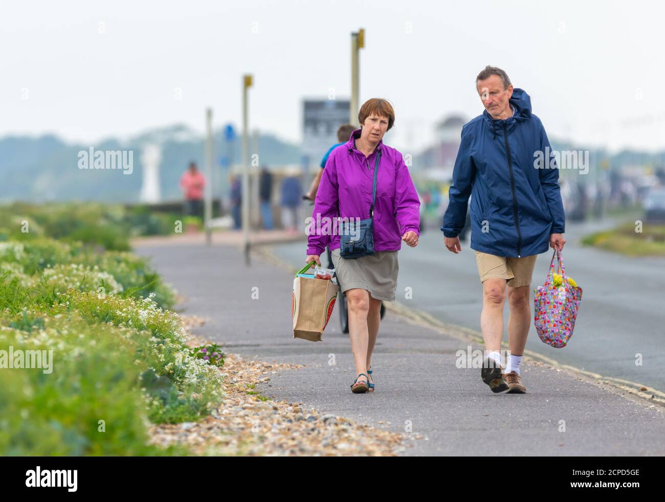 Coppia maschile e femminile di media età che cammina su un marciapiede indossando cappotti in una giornata fredda, portando pesanti borse per la spesa, nel Regno Unito. Foto Stock