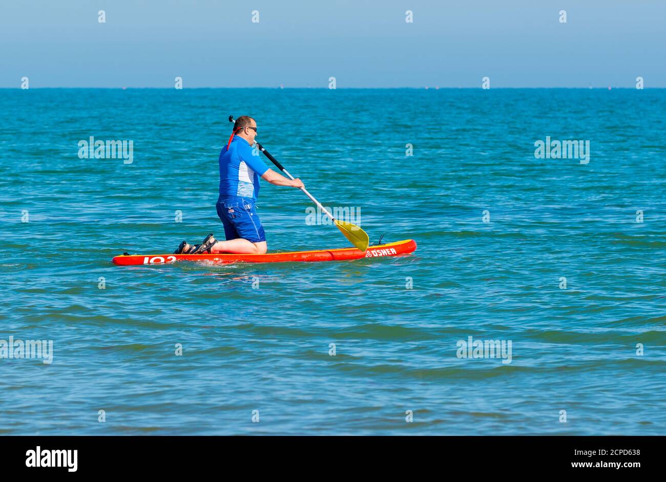 Un uomo inginocchiato su una tavola da paddle, paddle boarding in mare in una giornata calda in estate in Inghilterra, Regno Unito. Foto Stock