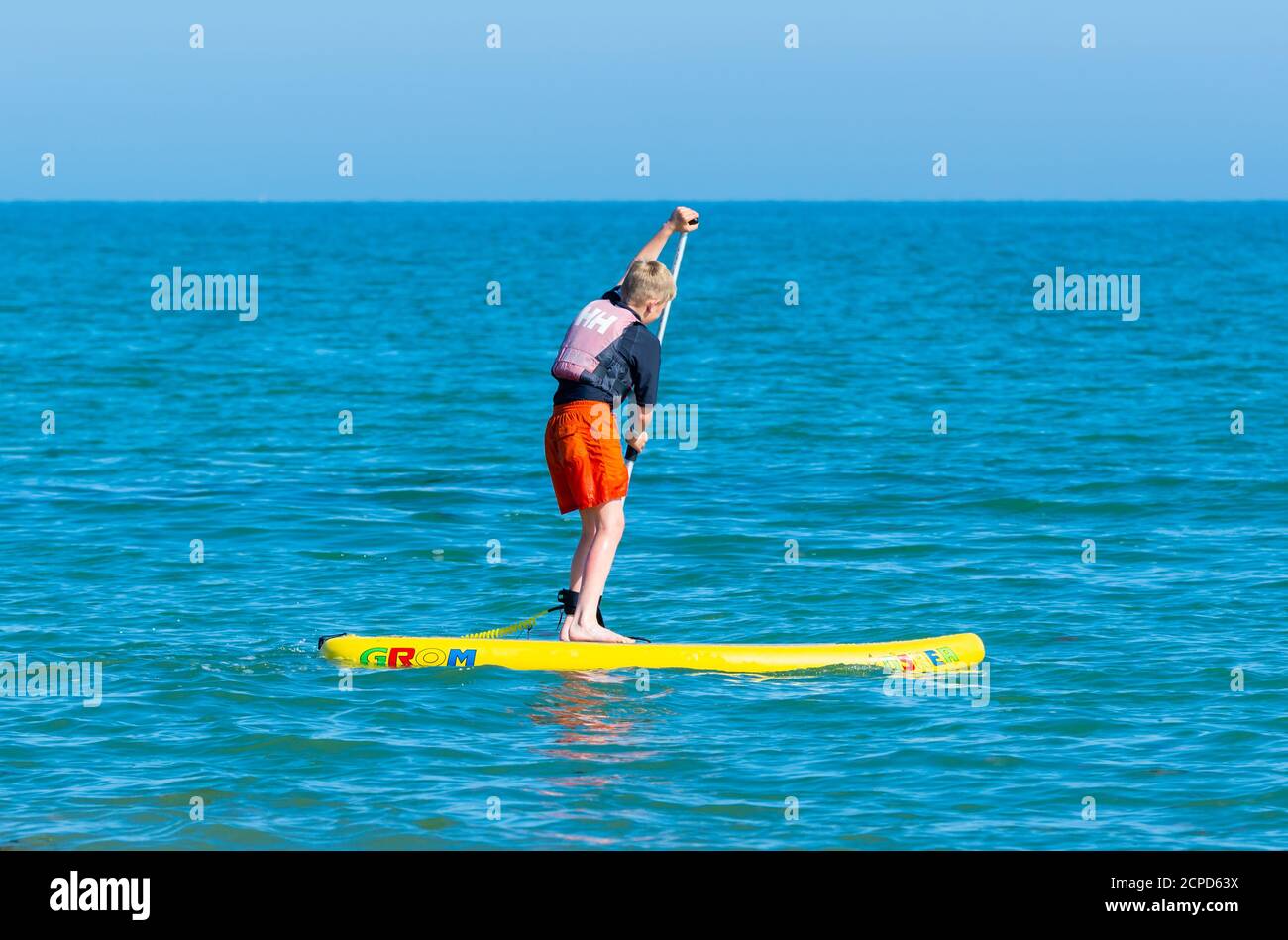 Un ragazzo in piedi su un paddleboard, paddle boarding in mare in una giornata calda in estate in Inghilterra, Regno Unito. Foto Stock