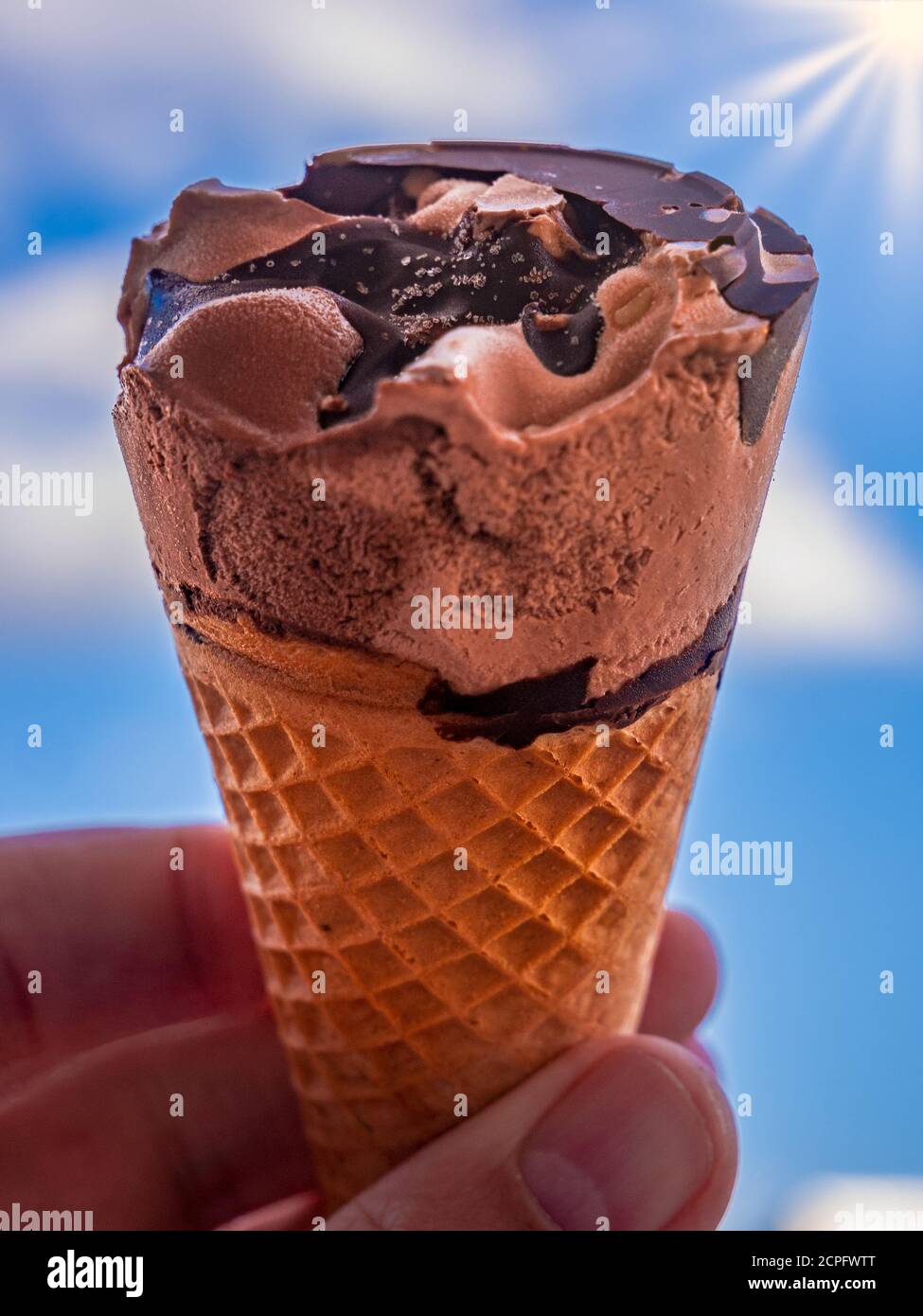 Cono gelato al cioccolato tenuto contro un cielo estivo con nuvole e sole con raggi nell'angolo in alto a destra Foto Stock