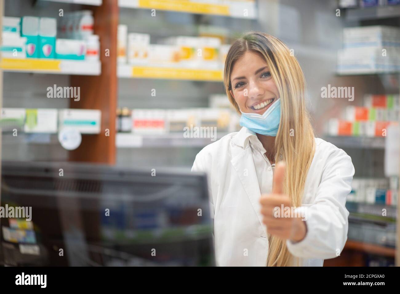 Ritratto di un giovane farmacista sorridente in una farmacia che dà pollici dietro un vetro, concetto di coronavirus Foto Stock
