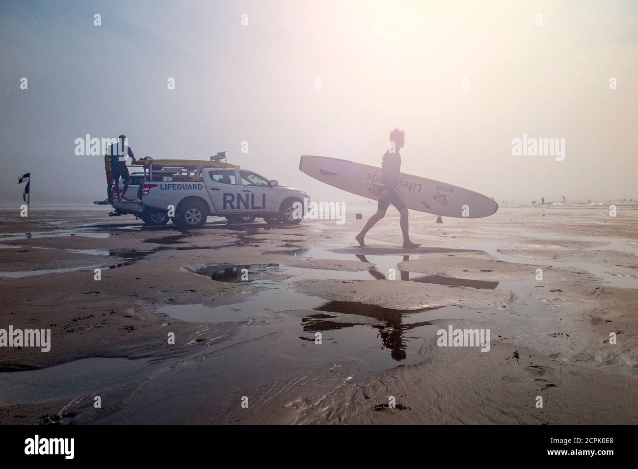 Un surfista che porta una tavola da surf vascola oltre la RNLI in pattuglia sulla spiaggia di Croyde, Devon Nord in alta estate Foto Stock