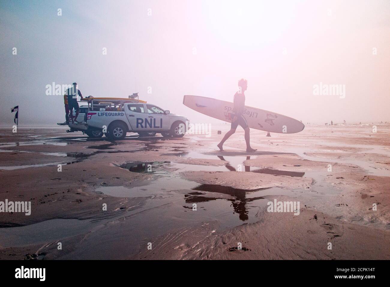 Un surfista che porta una tavola da surf vascola oltre la RNLI in pattuglia sulla spiaggia di Croyde, Devon Nord in alta estate Foto Stock