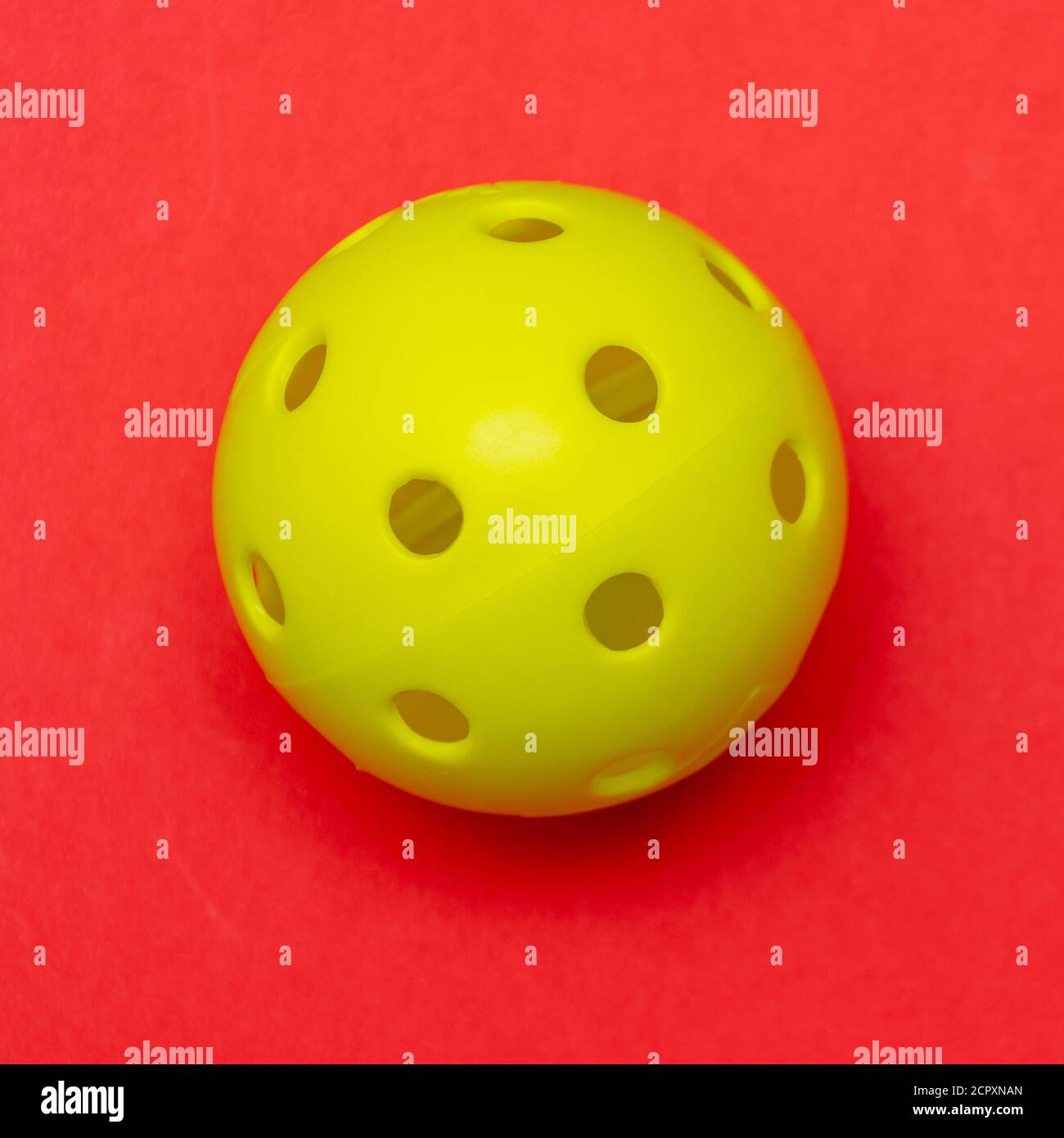 Di colore giallo brillante pickleball o sfera whiffle su un solido rosso luminoso appartamento sfondo laici che simboleggiano sport e attività con copia spazio. Foto Stock