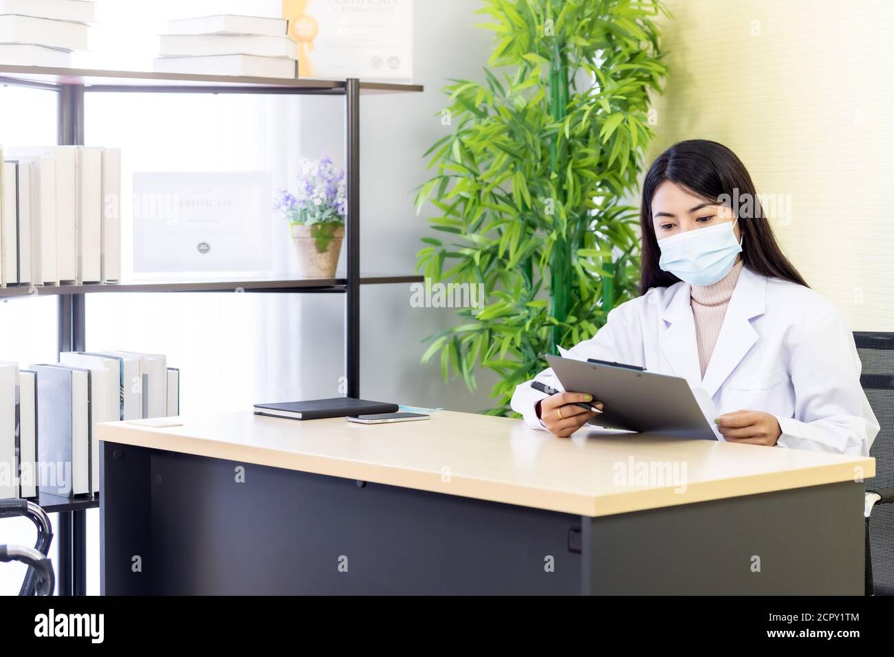 Ritratto di donna asiatica medico indossare maschera protettiva viso sedersi nella sua stanza di ufficio in clinica ospedale e leggere sul file di pazienza prima di controllare patie Foto Stock