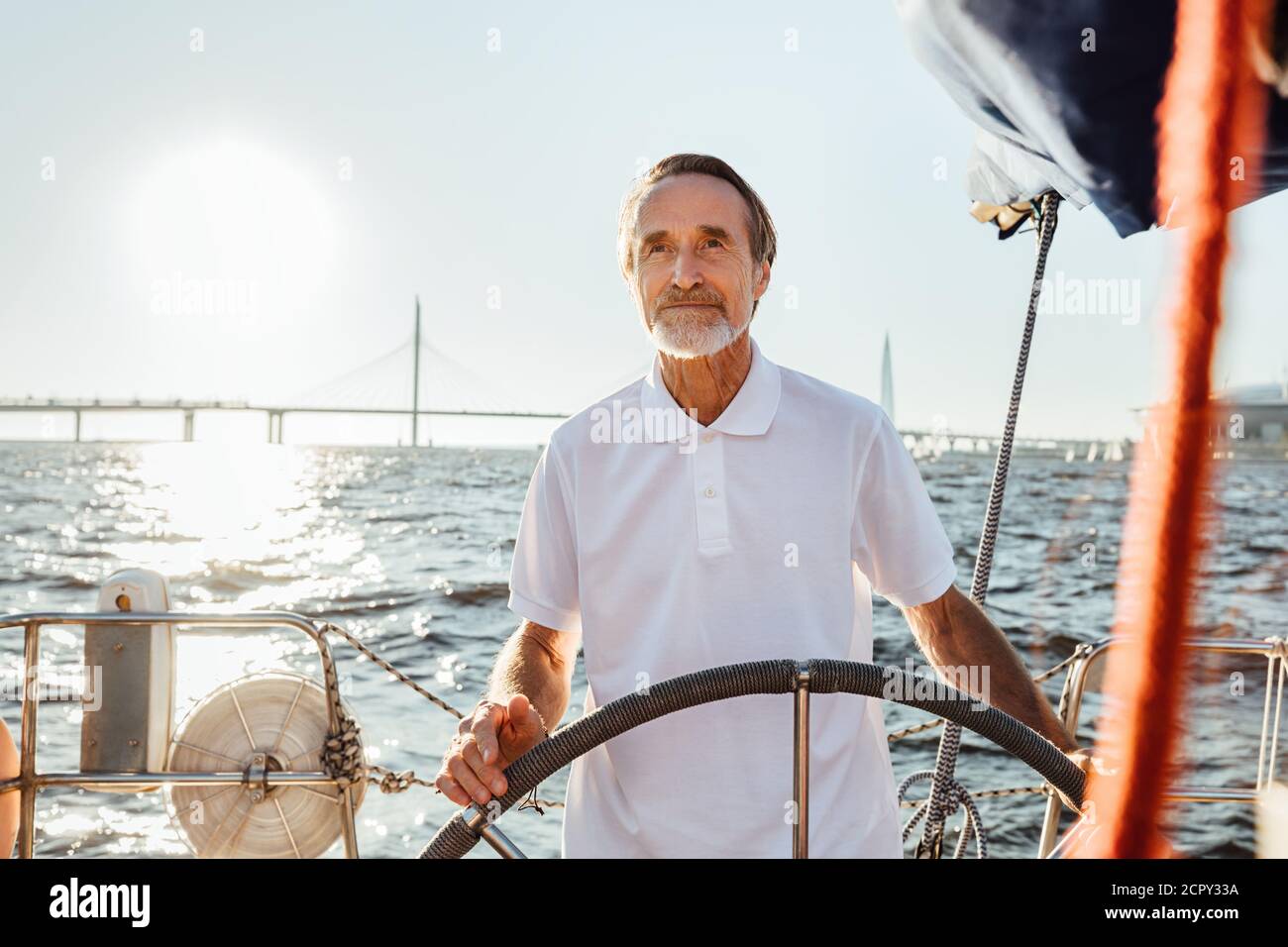 Uomo maturo che guida uno yacht privato mentre in vacanza. Yachtman bearded in piedi sulla sua barca a vela guardando a distanza. Foto Stock