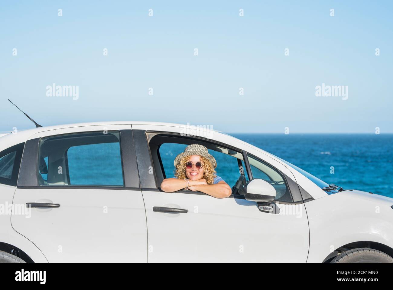 una bella donna felice guardando la macchina fotografica dall'interno di una macchina con il mare o l'oceano sullo sfondo - viaggiatore adulto e nomade stile di vita e. Foto Stock