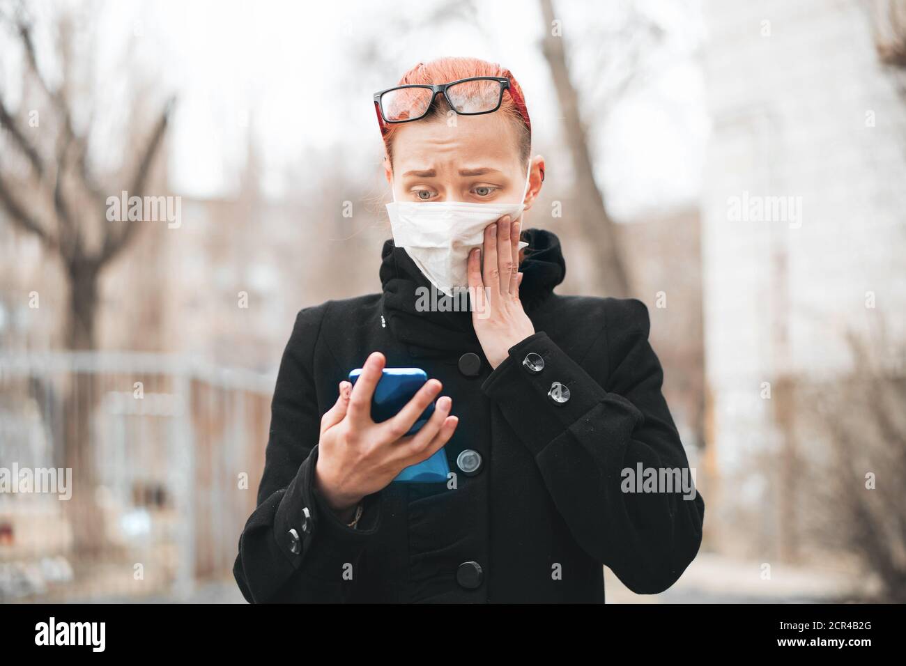 Zenzero ragazza in maschera guarda il telefono e legge notizie su epidemia. Protezione contro il concetto di virus. Foto Stock