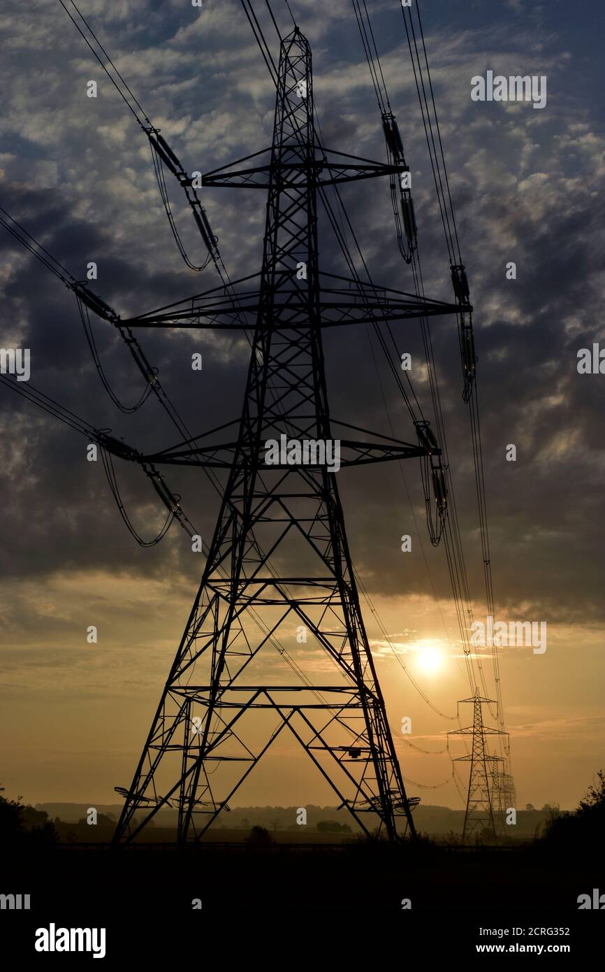 Silhouette della torre di trasmissione elettrica in pilone con alba e cavi elettrici che conducono a piloni in lontananza attraverso il paesaggio agricolo. Foto Stock