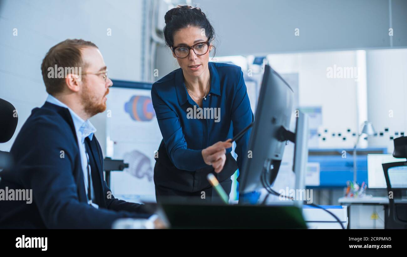 Modern Factory: La Supervisore del progetto femminile parla con un ingegnere industriale maschile che lavora su Personal computer. Utilizzano il software CAD per la progettazione e. Foto Stock