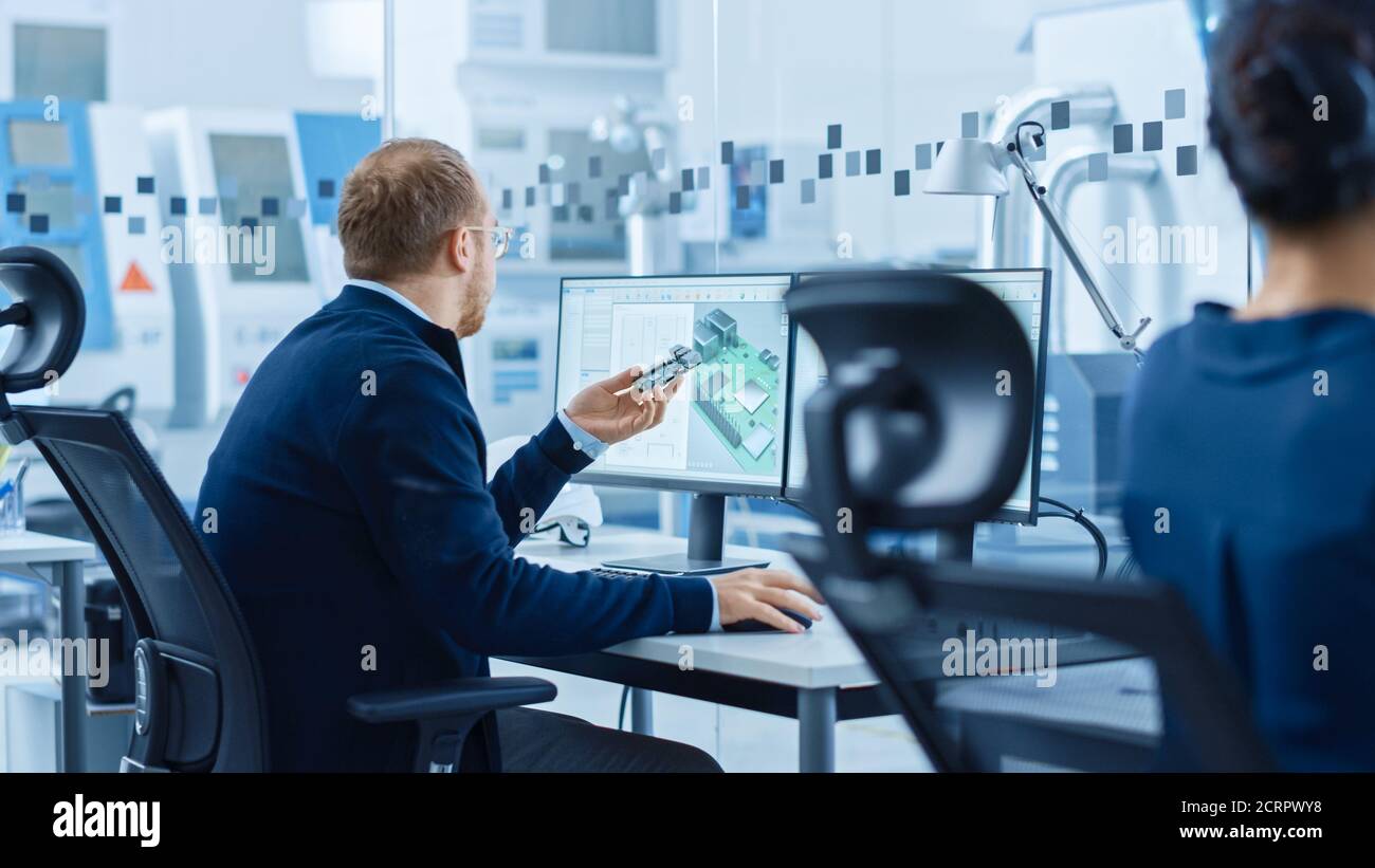 Electronics Factory: Male Electrical Engineer possiede un prototipo PCB, lavora su computer, utilizza il software CAD per sviluppare microchip, semiconduttori e. Foto Stock