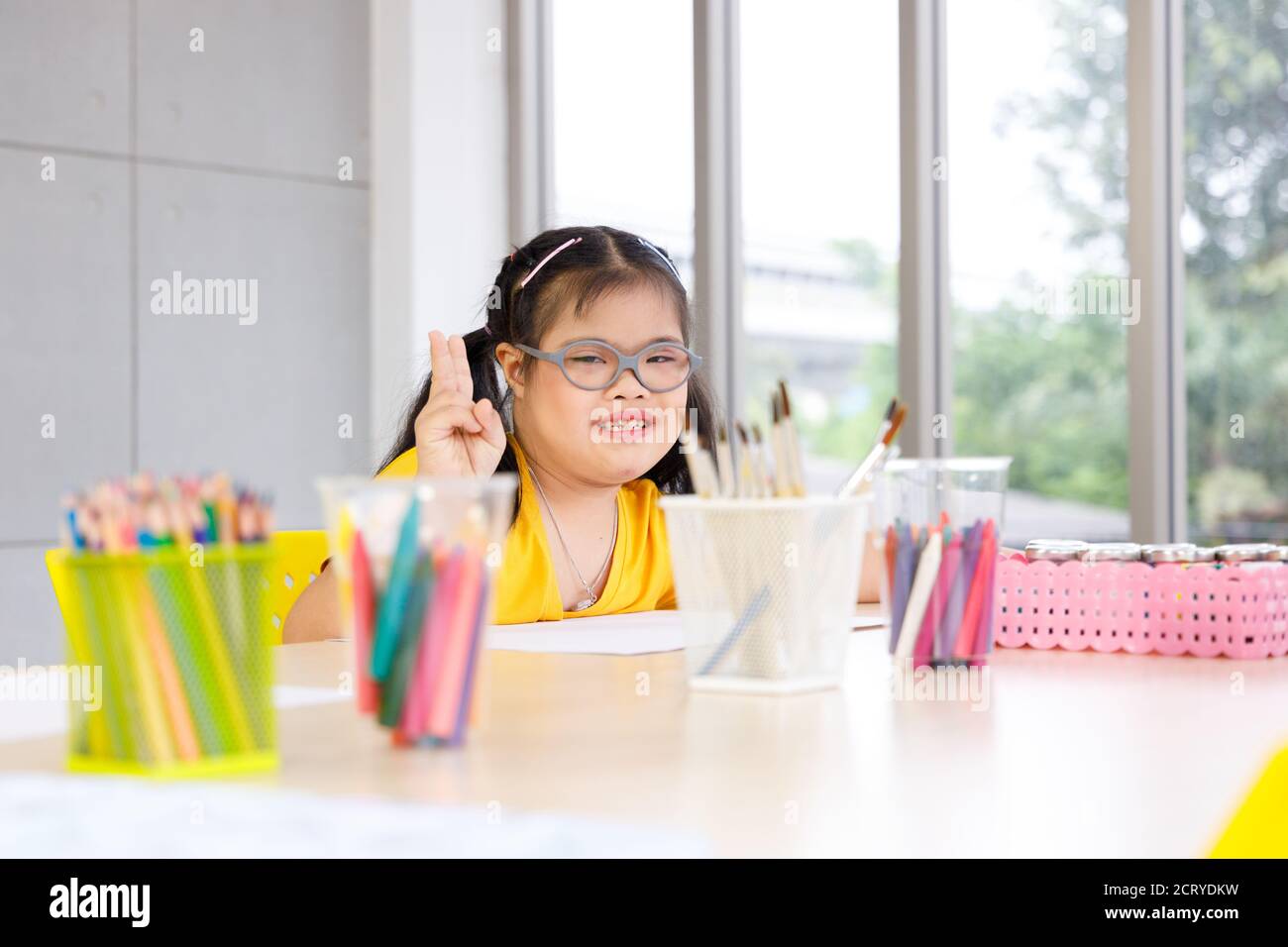 Ragazza asiatica felice con la sindrome di Down alzare due dita e sedersi alla scrivania piena di strumenti d'arte. Foto Stock